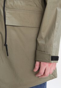 Gros plan sur une main à côté d'une manche de veste beige avec un poignet ajustable en Velcro et une grande poche avant avec rabat.