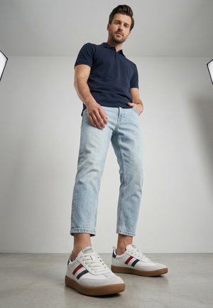 Mand i navy poloshirt og lyse jeans stående med afslappede hænder, iført hvide sneakers med røde og navy striber på gråt gulv.