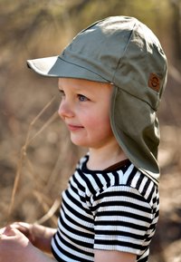 Světle olivově zelený sluneční klobouk se širokou krempou a klapkami na uši, s hnědou logo nášivkou. Dítě má na sobě černobíle pruhované tričko.