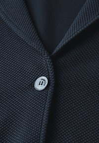 Tmavě námořnický texturovaný pletený cardigan s jedním bílým knoflíkem označeným "StreetOne" upevněným blízko límce.