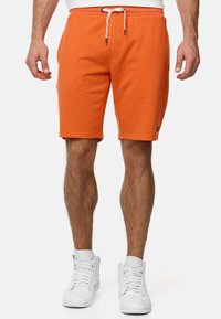 Mand iført orange knæ-lange shorts med snor og hvide høj-ankel sneakers, stående foran en helt hvid baggrund.