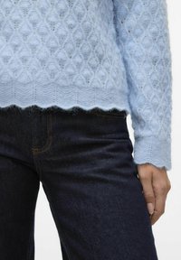 Lyseblå strikkegenser med diamantmønster, bølgekant og lett lodden tekstur, satt sammen med mørke jeans.