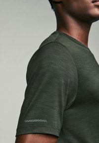 Dark green athletic shirt with short sleeves, featuring a subtle texture, crew neck, and a reflective accent near the hem of the sleeve.