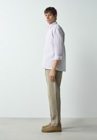 Junger Mann steht seitlich, trägt ein hell gestreiftes Hemd, beige Hose und hellbraune Wildleder-Sneaker vor einem schlichten hellen Hintergrund.