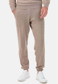 Pantalon de jogging marron clair en tissu doux. Dispose d'une taille élastique et de poignets élastiqués, avec des poches latérales. Porté avec des baskets blanches.