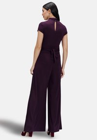 Purple jumpsuit with short sleeves, cinched waist, and tied back detail. Features a high neckline and wide leg design. Smooth fabric texture.