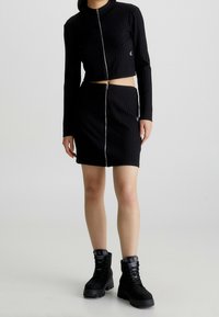 Chaqueta corta negra acanalada con cremallera frontal, combinada con una falda negra acanalada a juego con cremallera lateral, lucida con botas de combate negras.