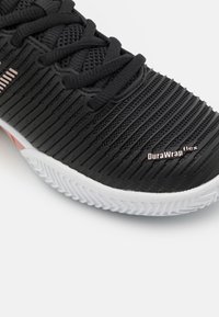 Črne atletske čevlje z teksturirano zgornjimi deli, ki vsebujejo vodoravne utore, ter belo gumijasto podplato. Opazno blagovno znamko označuje "DuraWrap flex."