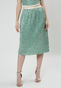 Falda verde esmeralda con lentejuelas, de longitud a media pantorrilla, diseño plisado y cinturilla color crema. Se lleva con sandalias plateadas de tiras.