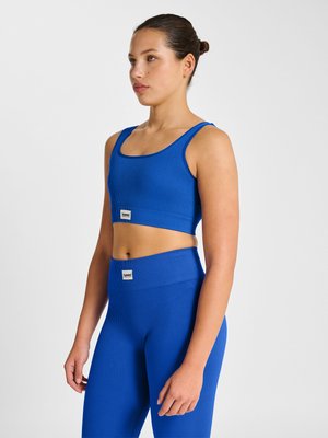 Jonge vrouw staat stil, draagt een blauw geribd crop top en hoog getailleerde sportleggings tegen een effen witte achtergrond.