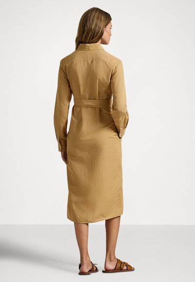 Kvinne som står, iført en beige midi-kjole med lang arm og midjebelte, samt brune flate sandaler, sett bakfra mot en hvit bakgrunn.
