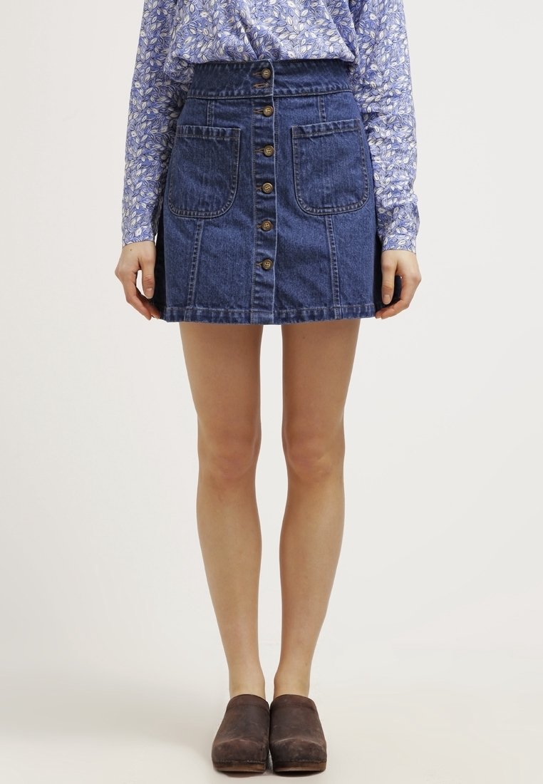 Jupe en denim avec un devant à boutons, deux grandes poches, une taille ajustée et un lavage bleu. Portée avec une chemise à manches longues à motifs.