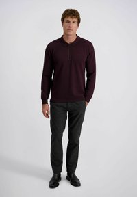 Homme portant un polo à manches longues bordeaux foncé, un pantalon gris foncé et des chaussures en cuir noires, se tenant contre un fond blanc uni.