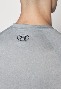 Camisa deportiva gris con cuello redondo, textura suave y un logo de Under Armour en negro en la parte posterior, cerca del escote.