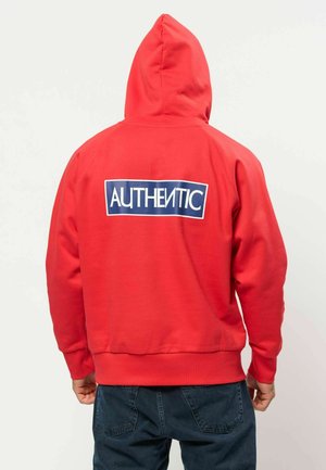 Persona que lleva una sudadera roja con capucha y un rectángulo azul en la espalda que muestra la palabra "AUTHENTIC" en letras blancas.