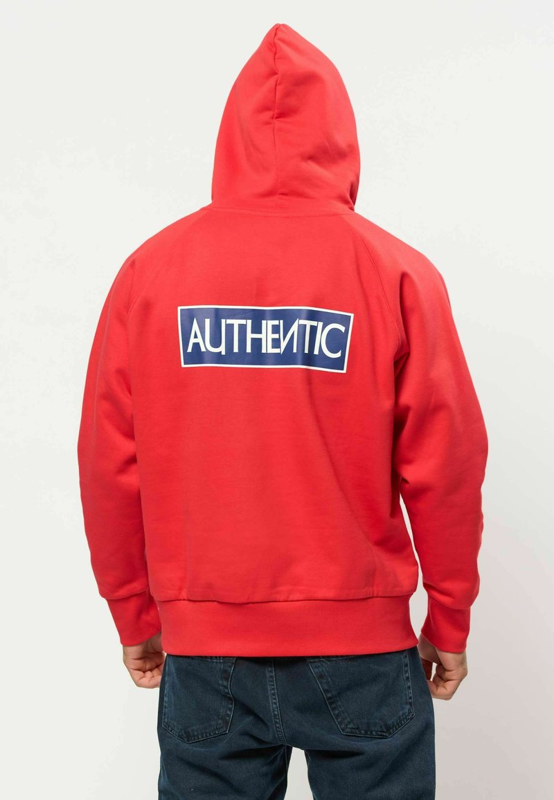 Personne portant un sweat à capuche rouge avec un rectangle bleu dans le dos affichant le mot "AUTHENTIC" en lettres blanches.