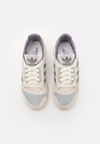 Adidas-sneakers med en ljusgrå och krämfärgad ovansida, med nät- och mocka-textur, tre mörkgrå ränder och vita snören.