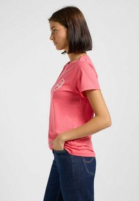 T-shirt en coton rose à manches courtes, col rond, avec un graphique de cœur blanc sur la poitrine, associé à un jean bleu marine.