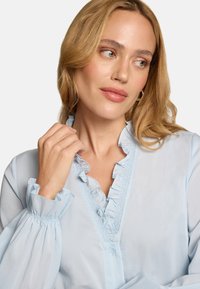 Blouse bleu clair avec poignets et col à volants, dotée d'une fermeture à boutons et de détails texturés le long de la patte de boutonnage. Tissu lisse.
