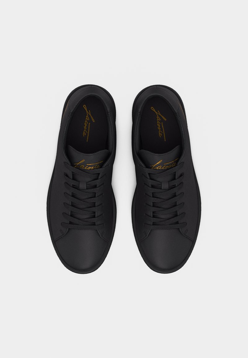 Een paar zwarte lage vetersneakers met afgeronde neuzen en een gouden geschreven logo op de binnenzolen, van bovenaf weergegeven op een witte achtergrond.