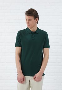 Polo-shirt vert foncé avec un tissu texturé, manches courtes, patte de boutonnage à trois boutons et col standard. Porté avec un pantalon de couleur claire.
