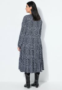 Musta mekko valkoisella sydänkuviolla, pitkät hihat, kerroksellinen malli ja röyhelöinen helma. Yhdistetty mustiin niittisaappaisiin. Näkyy takaa.