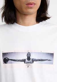 Hvit t-skjorte med et sentralt grafisk motiv av en basketballspiller med utstrakte armer, aksentuert med ordet "WINGS" over og en Nike-logo under.
