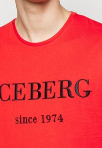Camiseta de algodón roja con cuello redondo, que presenta el texto negro en negrita "ICEBERG" y "desde 1974" debajo. Textura suave, ajuste informal.