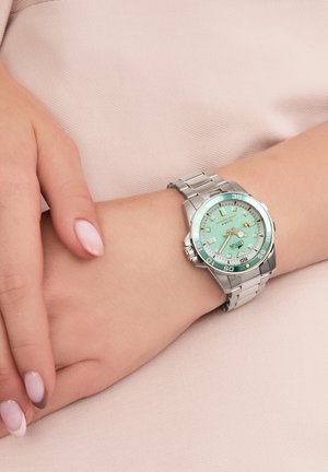 Reloj de acero inoxidable plateado con una esfera verde menta, marcaciones negras audaces y una pulsera suave, situado sobre un fondo rosa claro.