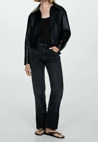 Chaqueta de cuero sintético negra con cuello, combinada con jeans oscuros de tiro alto con un dobladillo sin terminar y sandalias negras. Diseño sencillo.