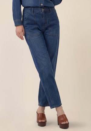 Frau trägt hoch taillierte blaue Jeans mit vorderen aufgesetzten Taschen und braunen Plateau-Sandaletten mit offenem Zehenbereich, steht mit überkreuzten Beinen.