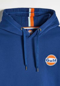Granatowy hoodie z regulowanym sznurkiem, z pomarańczowym i białym paskiem wewnątrz kaptura oraz okrągłym logo "Gulf" na klatce piersiowej.