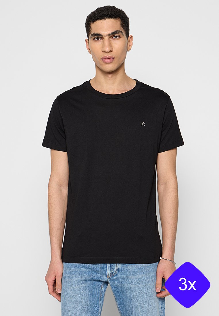 Junger Mann in einem einfarbigen schwarzen T-Shirt mit kurzen Ärmeln und hellblauen Jeans, der vor einem weißen Hintergrund steht.