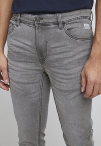 Szare jeansy z denimu o dopasowanym kroju, lekkim rozciąganiu, konstrukcji z pięcioma kieszeniami oraz małą naszywką z logo na pasku. Widoczna teksturowana powierzchnia.