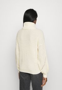 Cremefarbener, gestrickter Pullover mit hohem Kragen, gerippter Struktur und Ballonärmeln mit kleinen Ausschnittdetails.