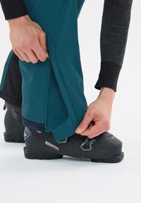 Teal utomhusbyxor med dragkedjeslutningar och justerbara remmar, i kombination med svarta skidstövlar som har strukturerade ytor och metalldetaljer.