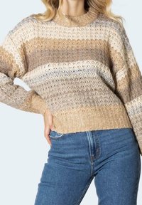 Femme portant un pull en tricot beige, crème et gris à rayures avec un motif texturé et un jean bleu taille haute, main posée sur la hanche.