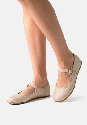Zapatos tipo Mary Jane de tela beige con hebilla dorada en los pies, suela plana y punta redondeada, usados por una persona de pie sobre un fondo claro.