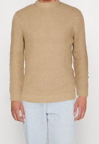 Jersey de punto beige con textura acanalada, cuello redondo y mangas largas. El dobladillo y los puños tienen un diseño acanalado que complementa el conjunto.