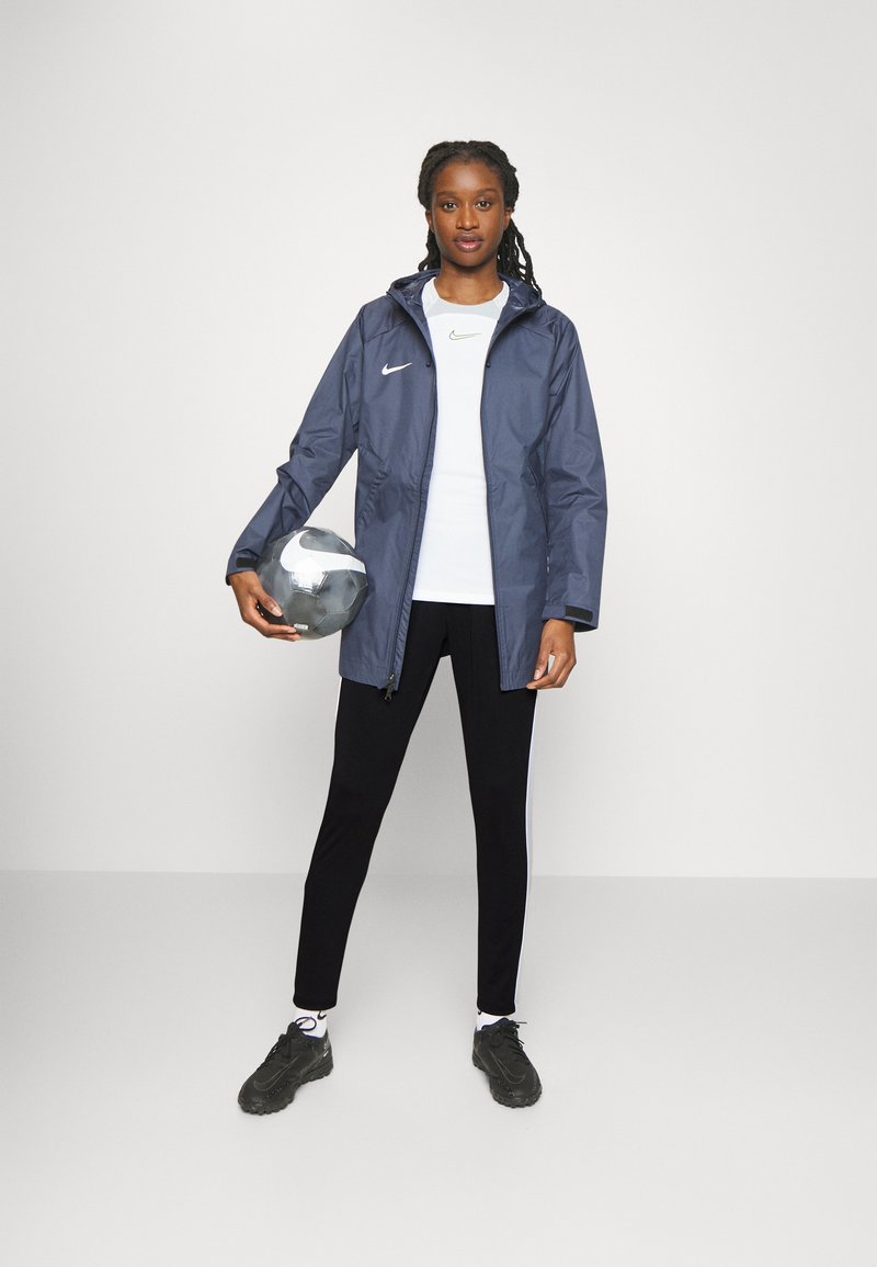 Marine waterdichte jas met capuchon, wit Nike-logo op de borst, gecombineerd met een witte top en zwarte sportbroeken, terwijl hij een zilveren voetbal vasthoudt.