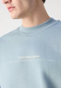 Gaiši zils sudraba krekls ar ribinātu apkakli, uz kura ir balts uzraksts "Calvin Klein Jeans" un "IZVEIDOTS 1978. GADĀ."