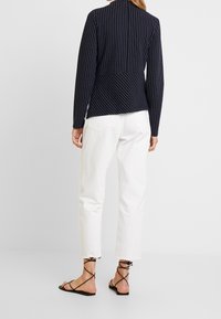 Marinblå kavaj med vertikala vita ränder, kombinerad med vita cropped-byxor och svarta remsandaler, sedd bakifrån.