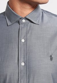 Šedá košile s herringbone vzorem, klasickým límcem, bílými knoflíky a malým vyšitým logem na hrudi. Měkká textura látky.