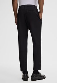 Schwarze Hose mit schmal zulaufendem Schnitt, elastischem Bund und zwei Gesäßtaschen. Kombiniert mit formellen schwarzen Schuhen und Socken. Glatte Stoffoberfläche.