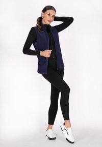 Marineblaues ärmelloses Vest mit hohem Kragen und Reißverschluss, kombiniert mit schwarzen langen Leggings und weißen Sneakers mit schwarzen Akzenten.