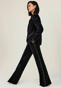 Vrouw in een zwart fluwelen tuxedo met een satijnen streep langs de broek, met handen in de zakken, lopend tegen een eenvoudige neutrale achtergrond.