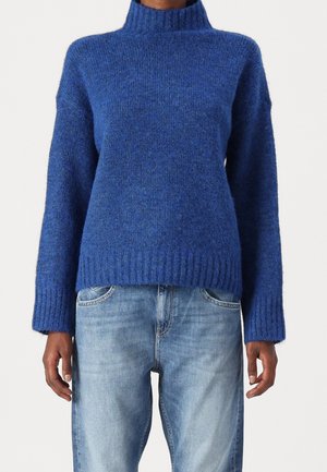Blå, fuzzy strikket sweater med høj rib-krave og lange ærmer, parret med lyseblå jeans. Sweateren har en afslappet pasform.