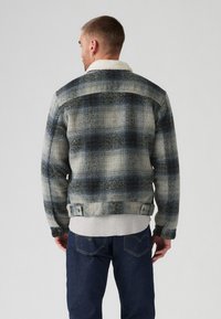 Chaqueta de cuadros en gris y azul con cuello de borreguillo. Diseño ajustado, tela texturizada y puños con botones. Llevada sobre un suéter gris.