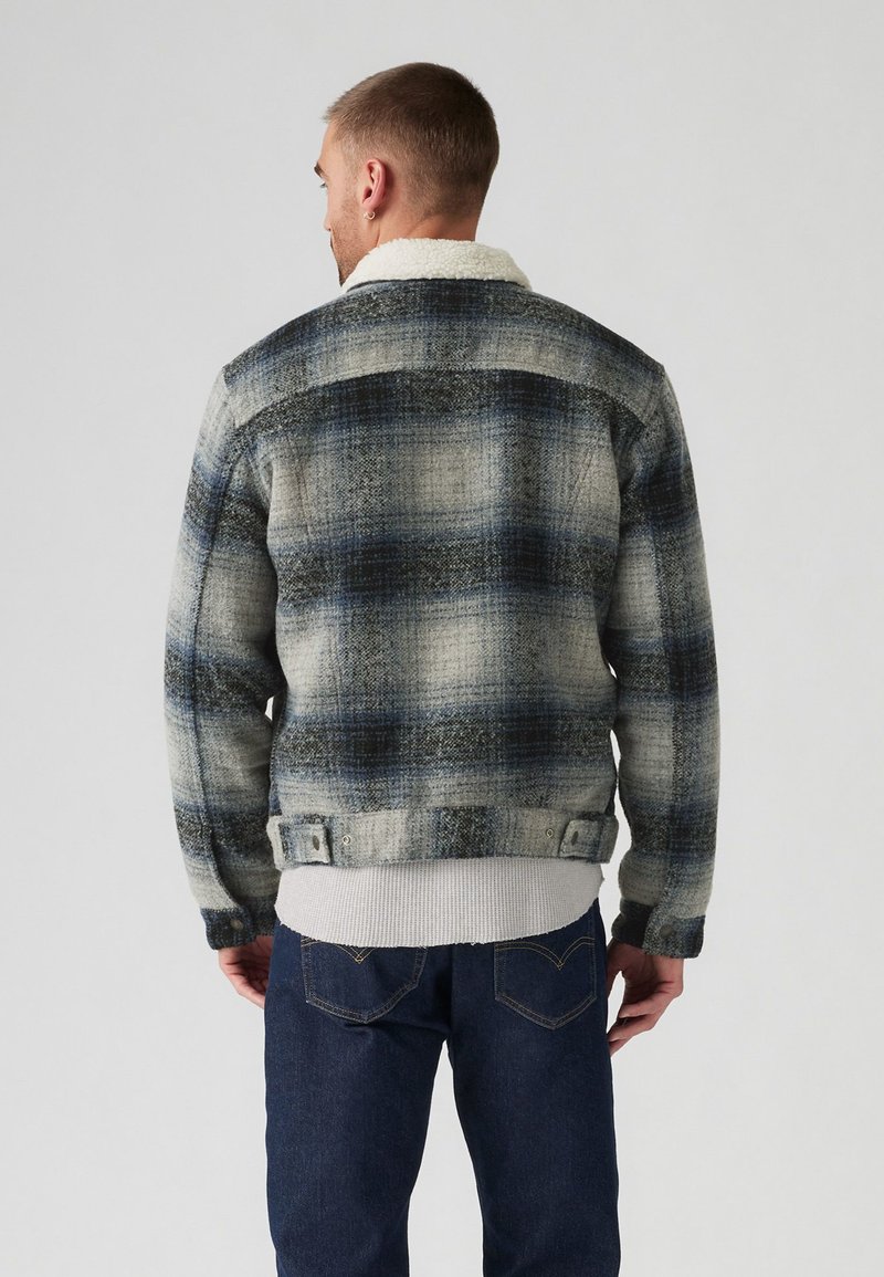Veste à carreaux en gris et bleu avec un col en fausse fourrure. Design ajusté, tissu texturé et poignets boutonnés. Portée sur un pull gris.