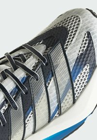 Sportska cipela s gornjim dijelom od mrežaste tkanine u crnoj, sivoj i plavoj boji, s teksturiranim sintetičkim umecima i bijelim slojem između potplata naglašenim plavim đonom.